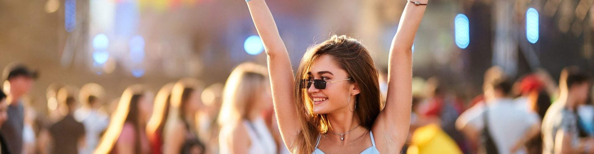 Arms raised, peace sign, dancing in crowd with stage lights. Happy young woman enjoys summer music festival on beach. Casual fashion, sunglasses, fun with friends at sunset concert party.